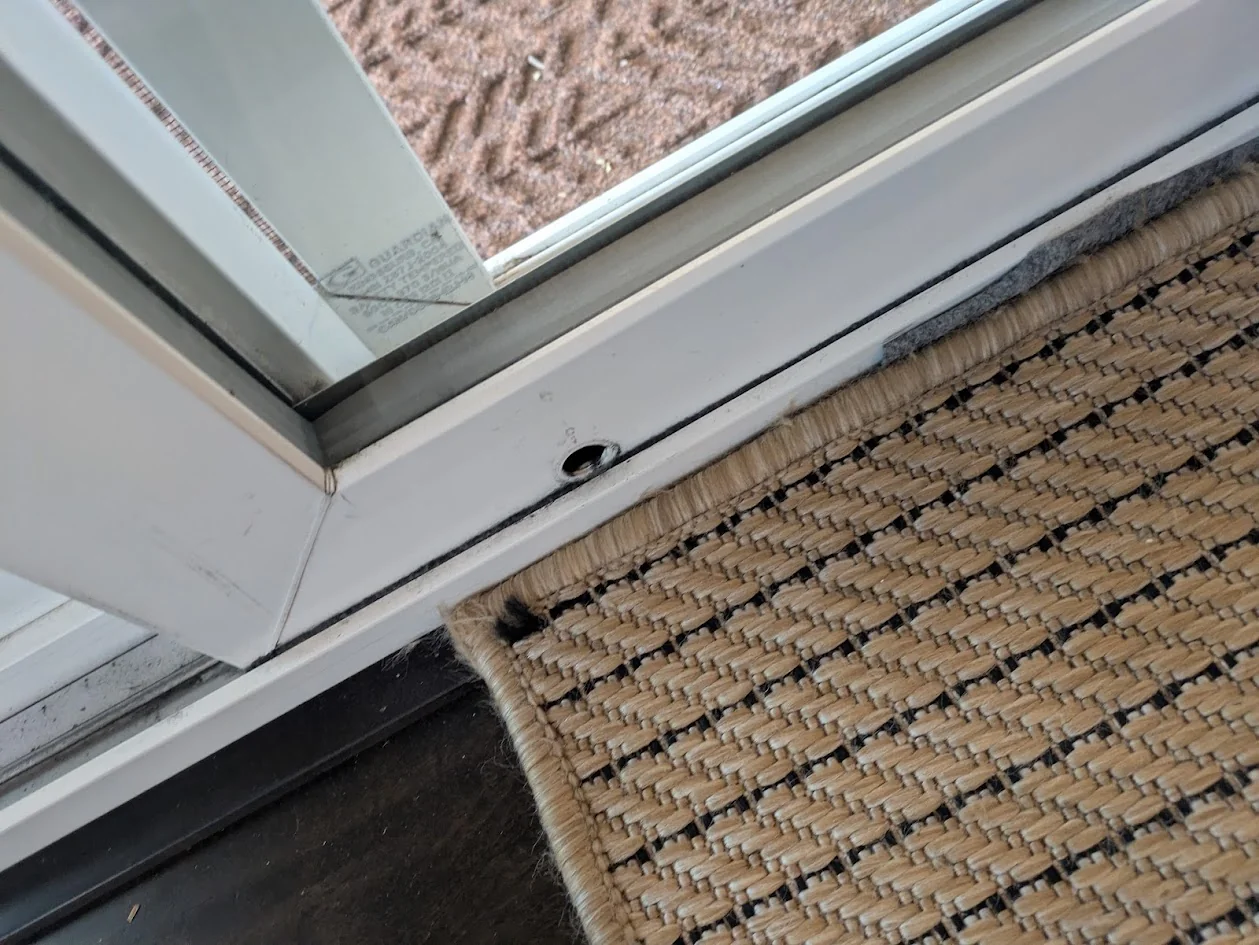 A close-up of a sliding patio door bottom edge showing the small roller adjustment screw hole near the corner
