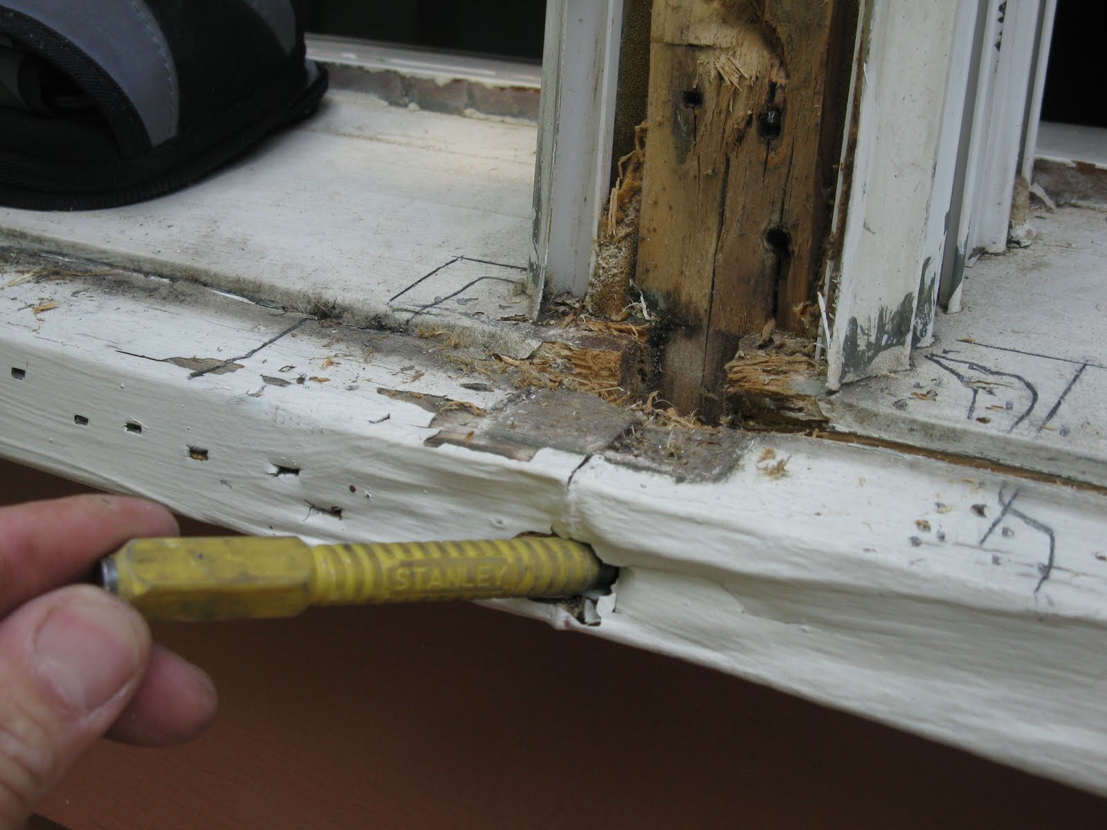 A close-up of a window sill corner being probed with a screwdriver, showing flaking paint and dark, softened wood near the edge