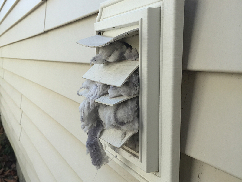 A close-up photo of a dryer vent duct connection with visible lint accumulation around the metal collar and inside the duct