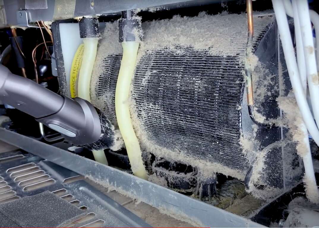 A closeup of dusty refrigerator condenser coils being cleaned with a coil brush and a vacuum crevice tool