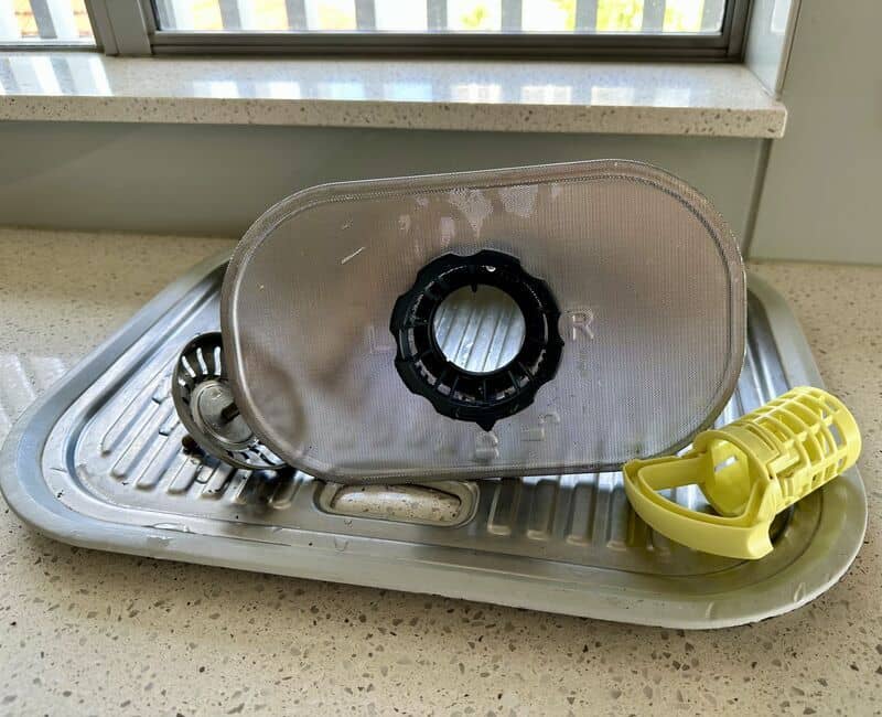 A cylindrical dishwasher filter and flat mesh screen sitting in a kitchen sink next to a nylon brush and dish soap