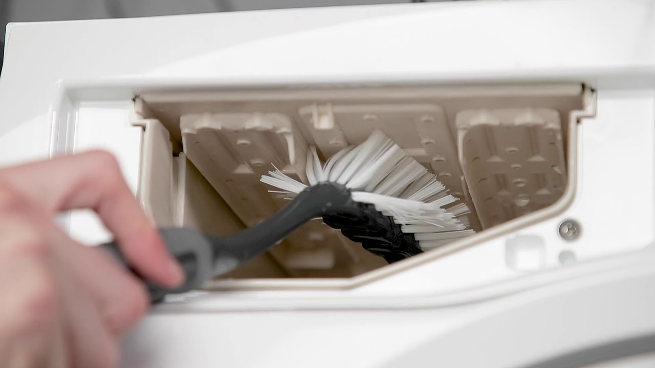 A detergent drawer from a front-load washer sitting in a sink while being scrubbed with a small brush