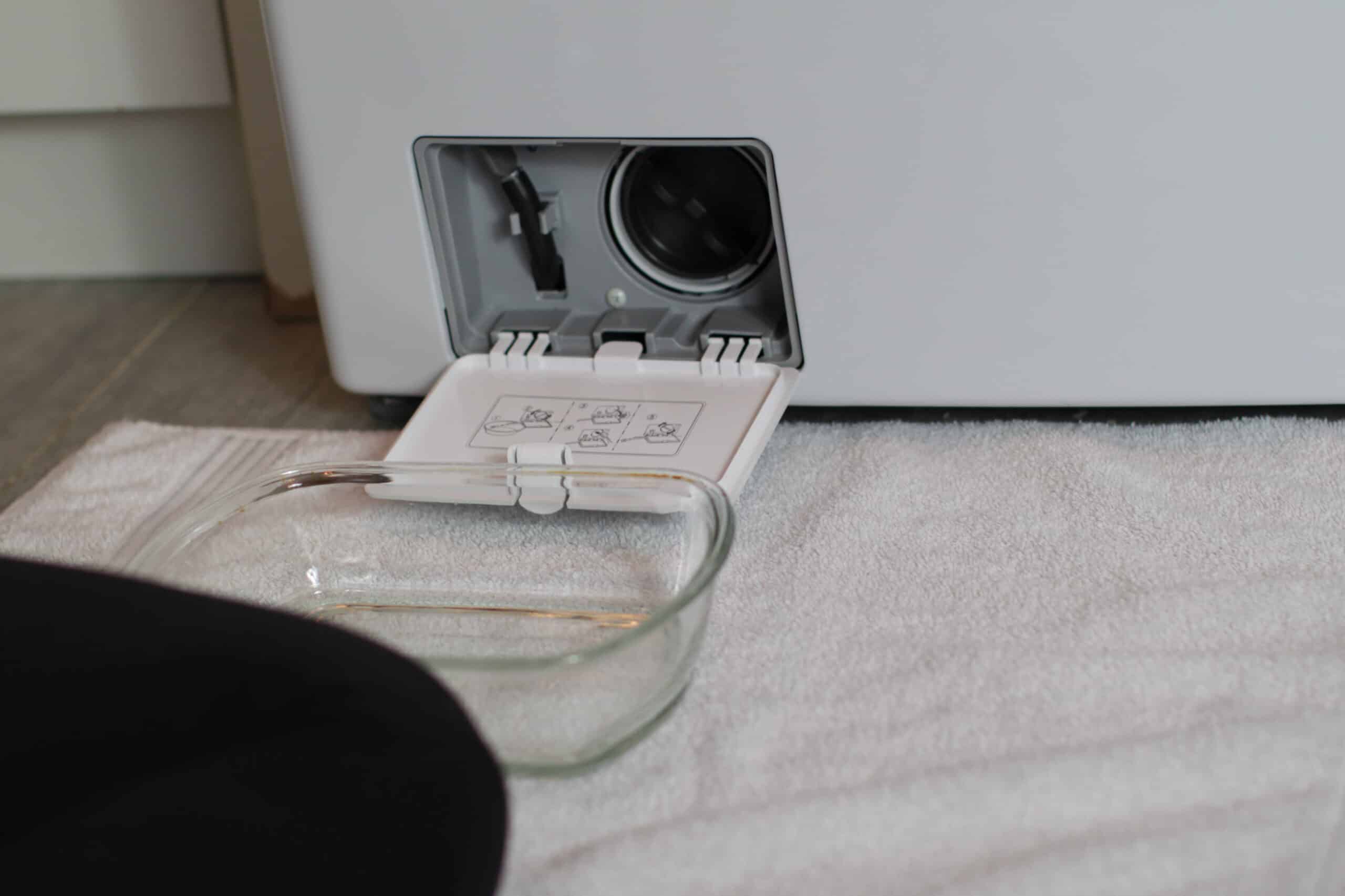 A front-load washer lower access door opened to show the drain pump filter cap being removed with towels placed underneath