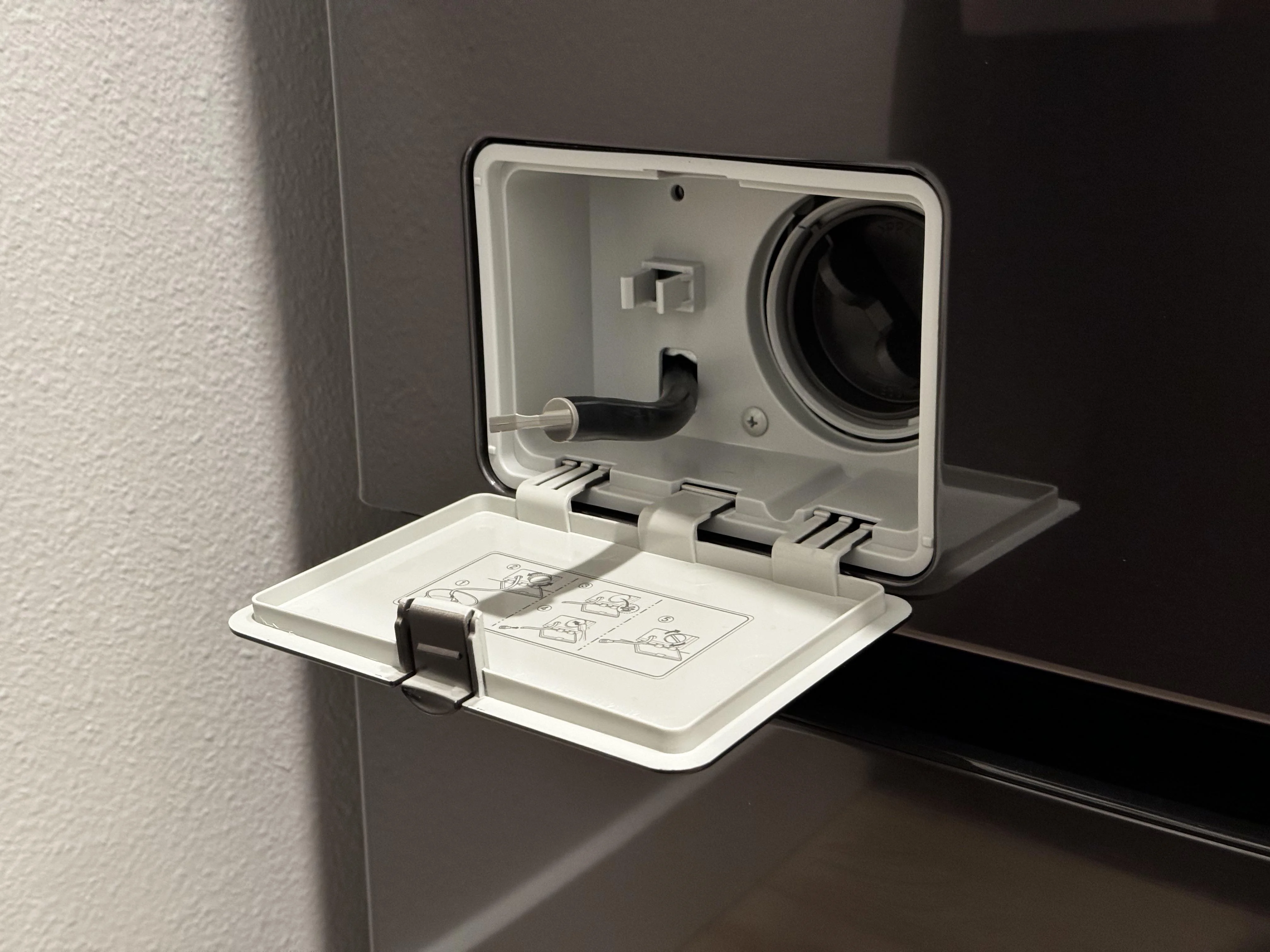 A front-load washer lower access panel open, showing the drain pump filter knob and a small emergency drain hose