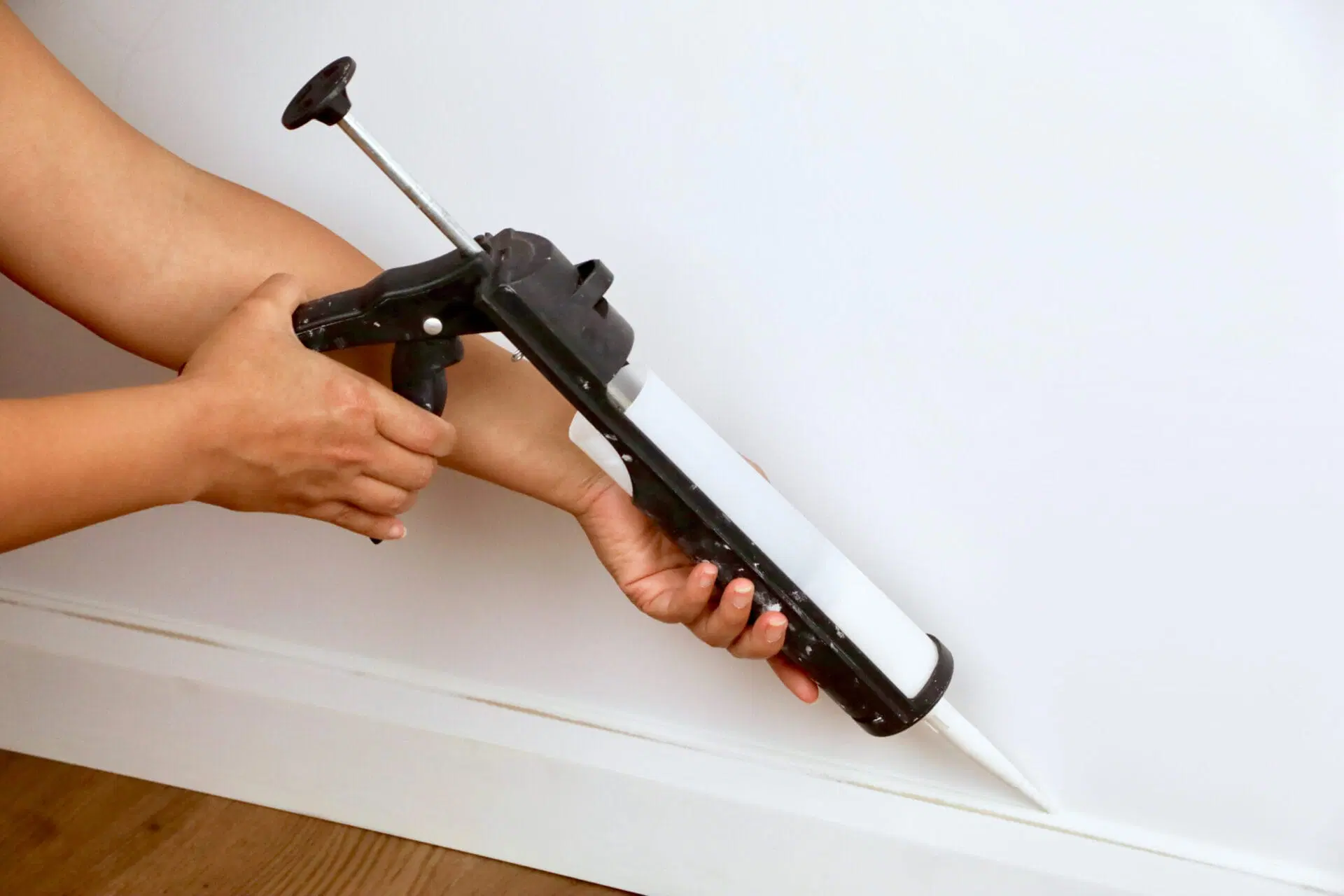 A hand applying a thin bead of paintable caulk along the seam between an old baseboard and a newly spliced patch piece