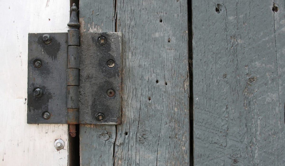 A hand rubbing a plain white candle along the seam of a metal door hinge on a painted door jamb