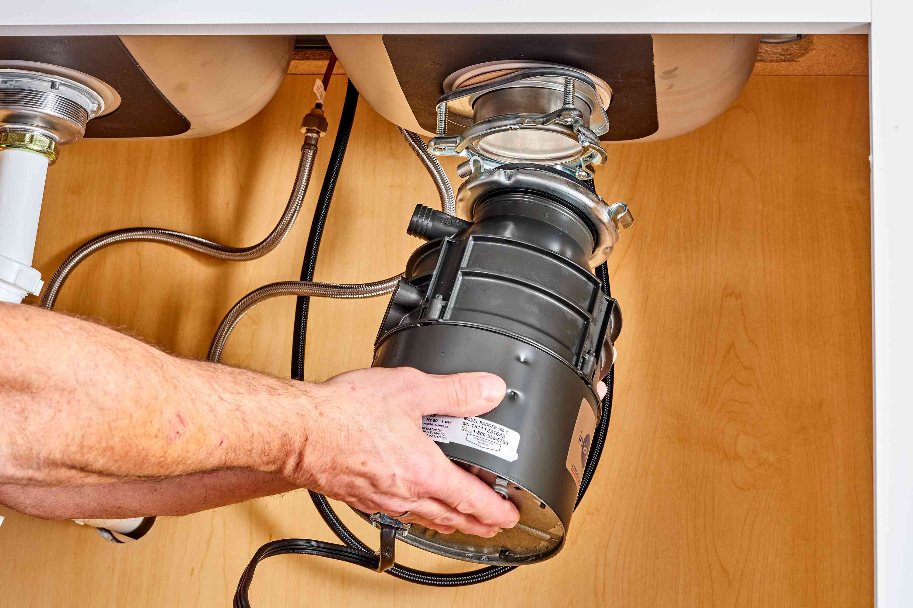 A homeowner using a flashlight to inspect a garbage disposal and drain connections under a kitchen sink while holding a paper towel near the mounting ring