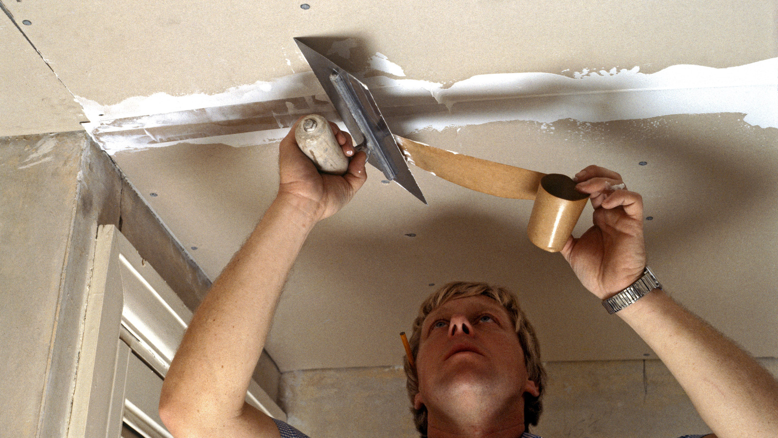 A person applying joint compound to a ceiling patch with a wide drywall knife, smoothing the edges into surrounding paint