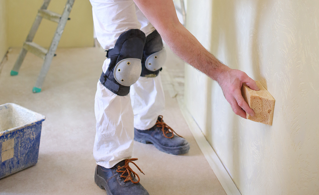 A person sanding a dried drywall patch with a sanding sponge while holding a shop vacuum hose nearby to capture dust
