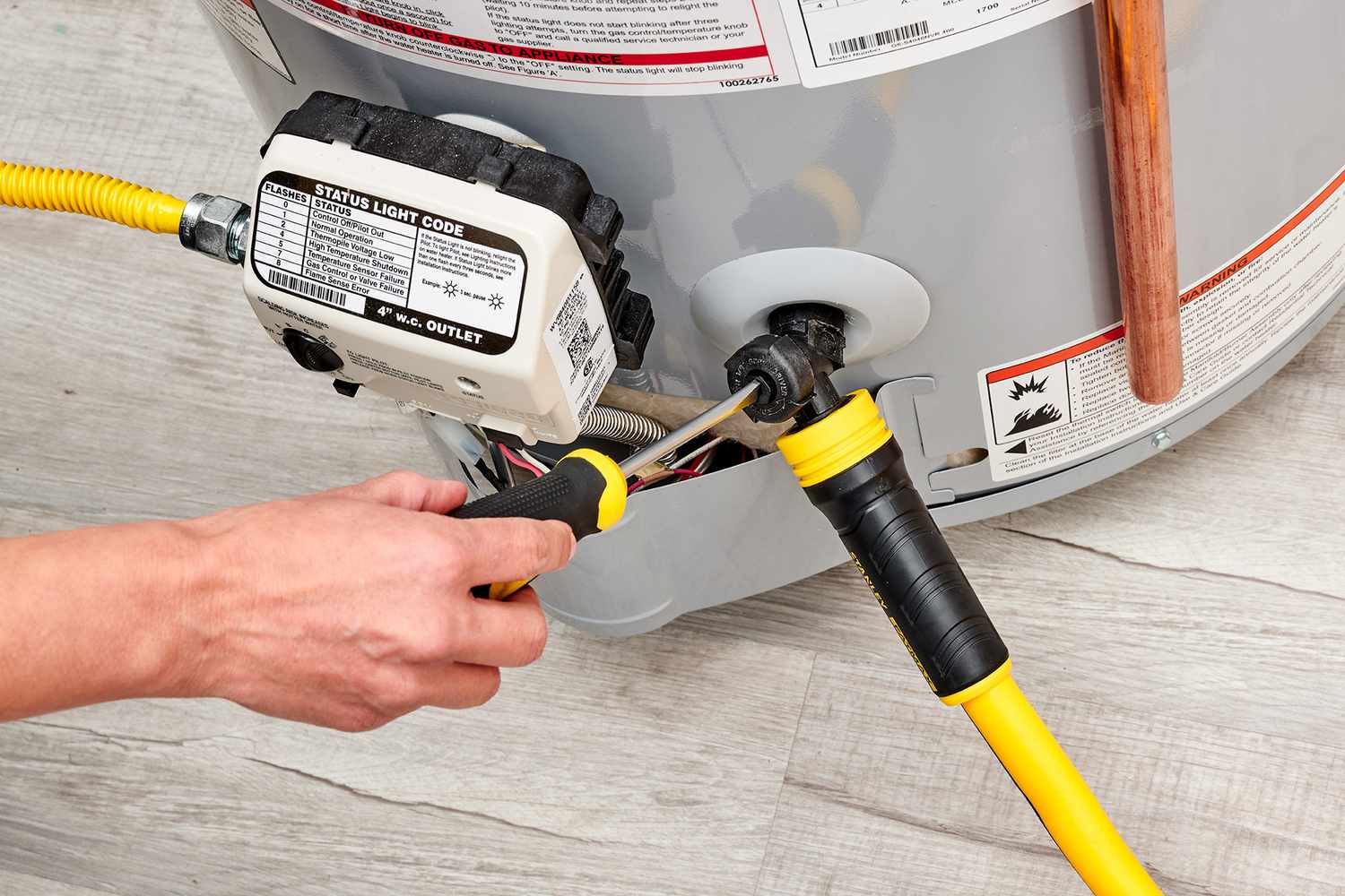 A residential tank-style water heater with a garden hose connected to the drain valve near the bottom, hose routed toward a floor drain