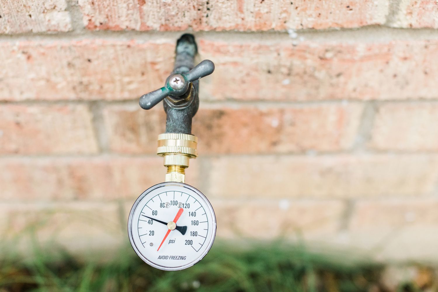 A round water pressure gauge threaded onto an outdoor hose spigot on the side of a house