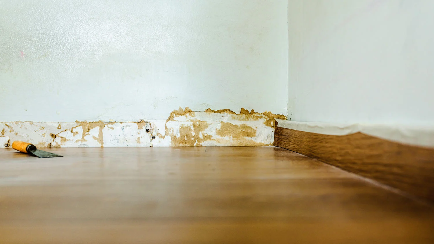 A small pry bar lifting a baseboard away from a wall while a putty knife protects the drywall surface