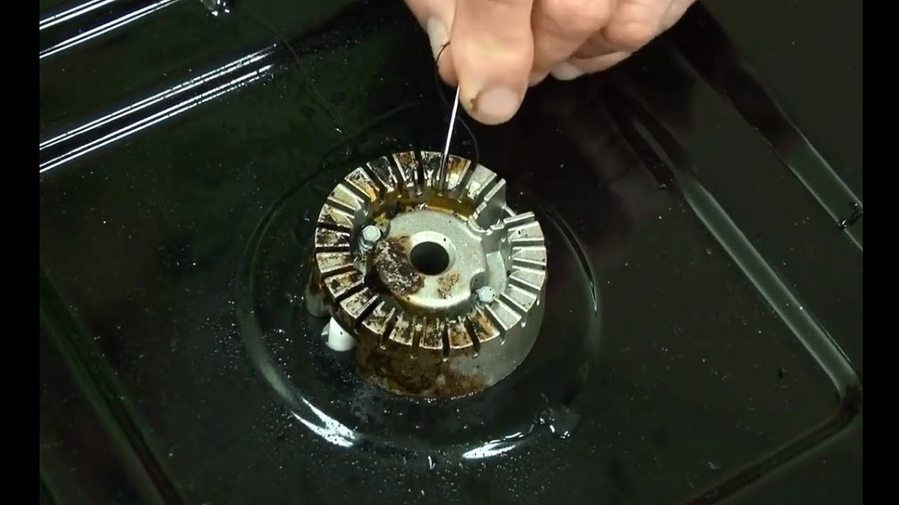 A toothbrush scrubbing cooked-on residue from a gas stove burner head over a kitchen sink