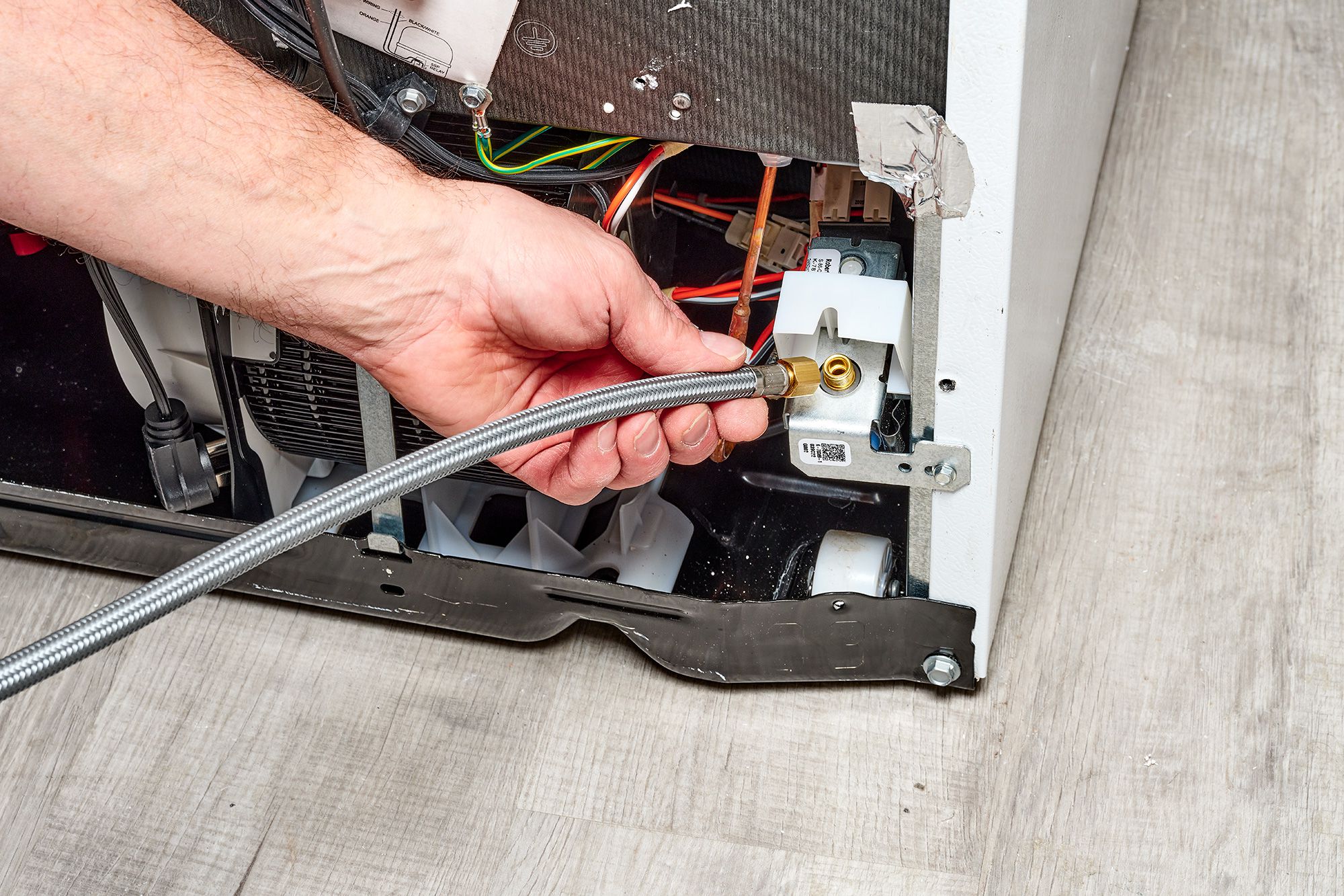 A view behind a refrigerator showing the braided water supply line connected to the fridge inlet valve