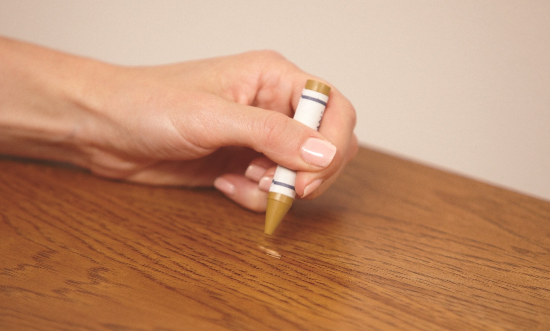 A wax fill stick and plastic scraper next to a hardwood floor scratch being filled with matching wax