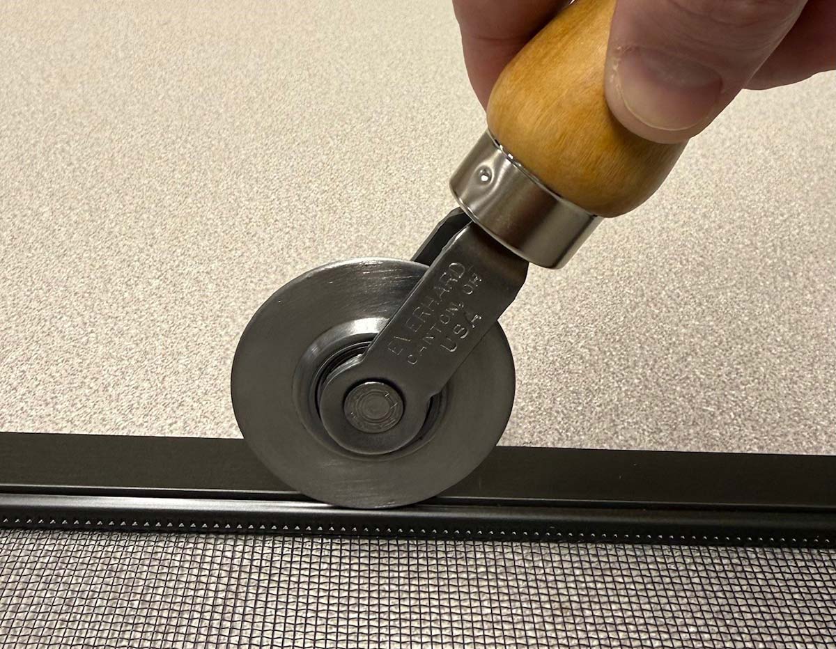 A window screen spline roller tool resting on a workbench next to a screen frame and a roll of replacement mesh