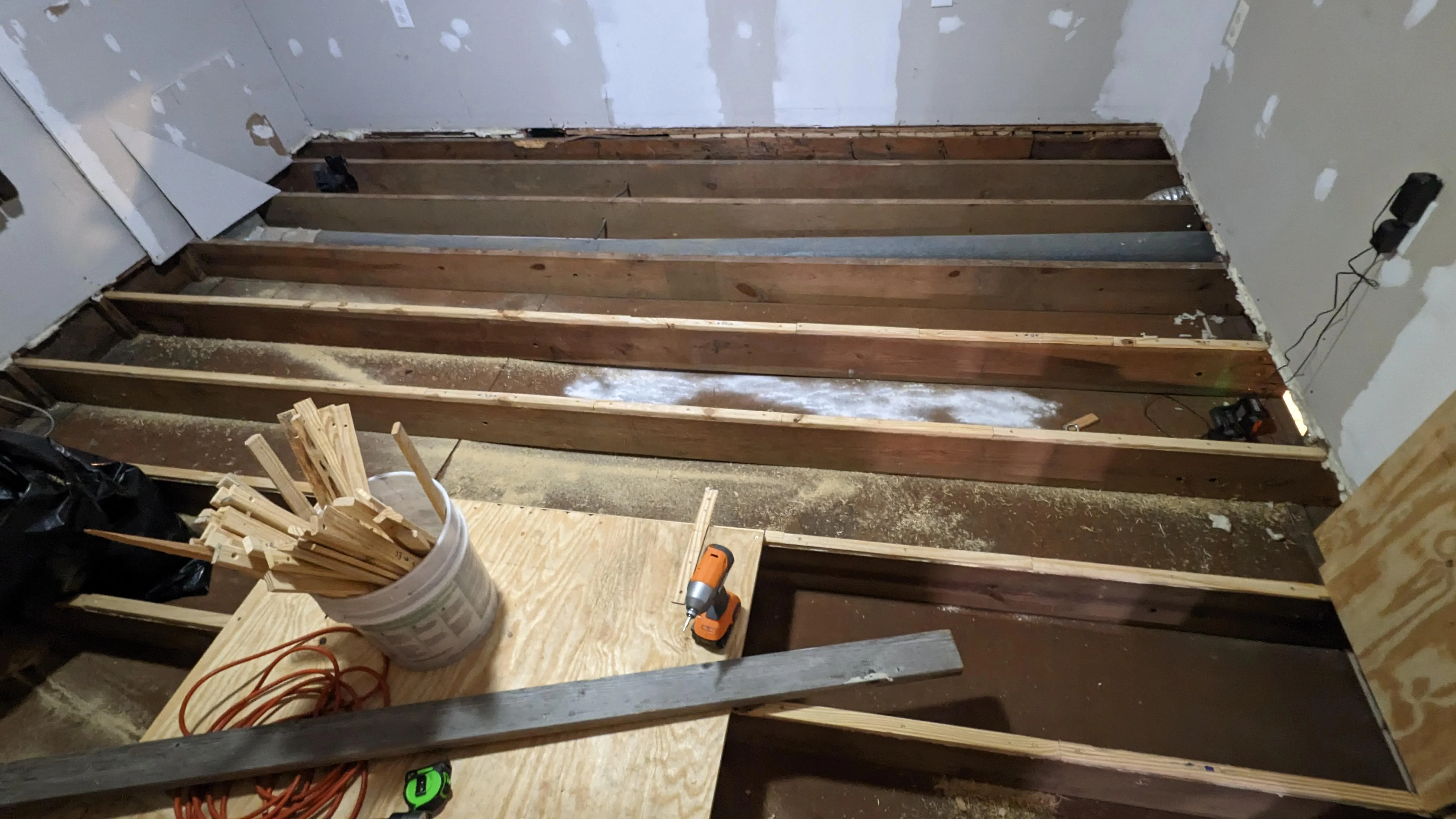 From a basement ceiling, a wood shim being tapped between a floor joist and the underside of a plywood subfloor