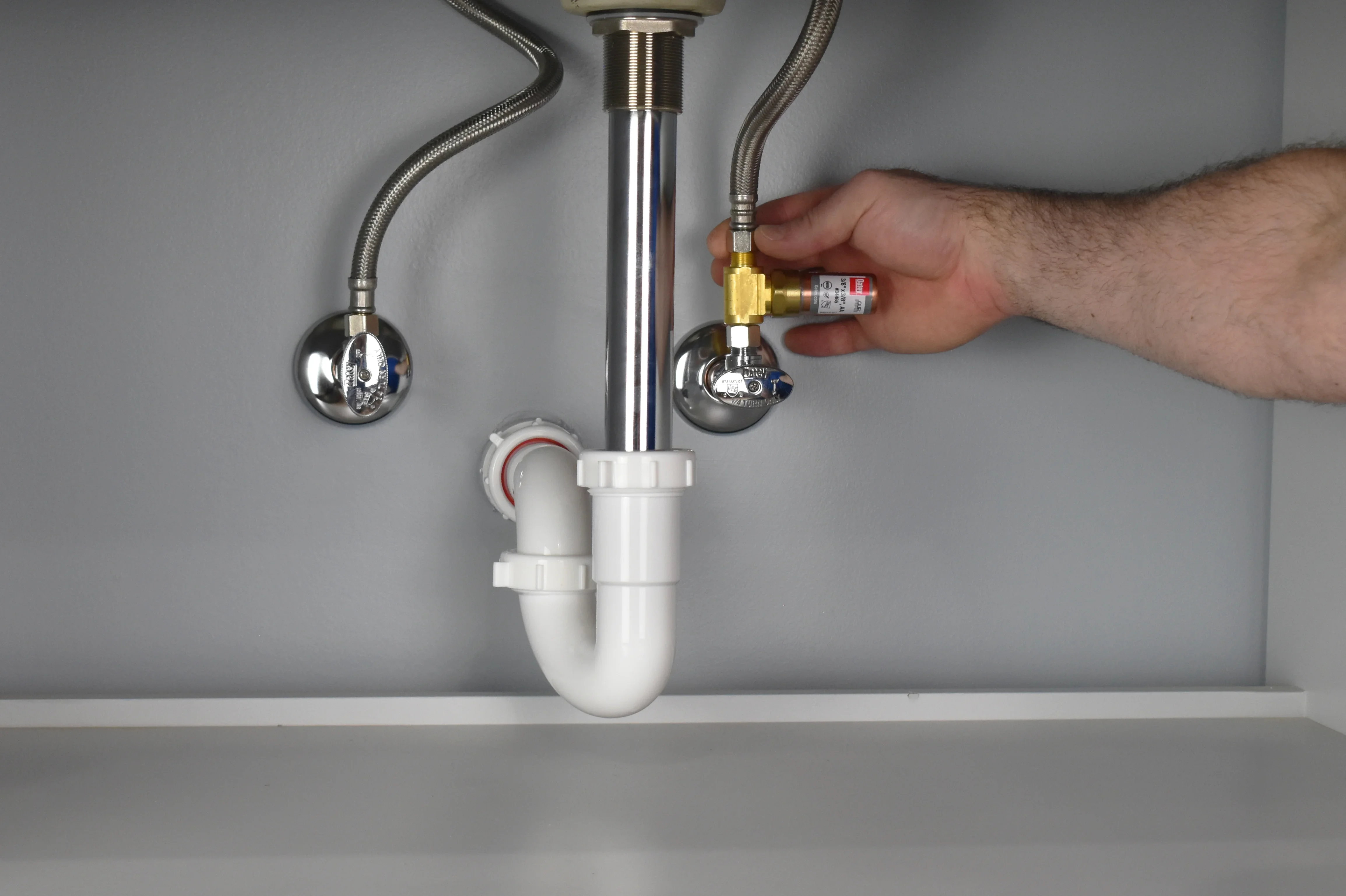 Under a bathroom sink, a small metal water hammer arrestor is attached at an angle stop valve on a braided supply line