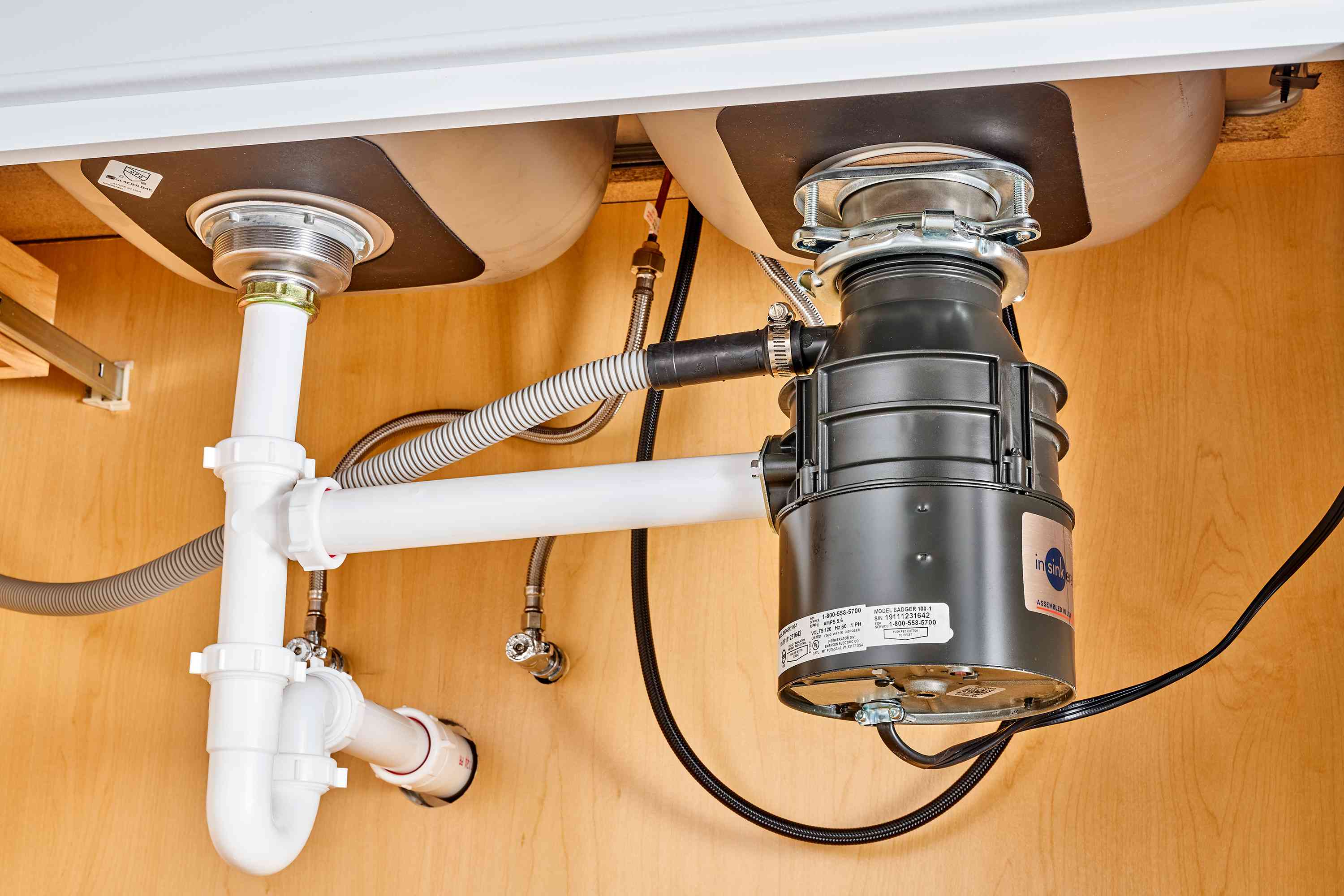 Under a kitchen sink, a garbage disposal outlet and discharge tube connection show moisture around the joint